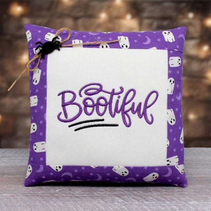 A square decorative throw pillow with a purple border patterned with white ghosts, moons, and stars, featuring a white center panel with the word 'Bootiful' embroidered in purple script. A small black spider decoration and twine are attached to the top left corner.