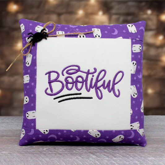 A square decorative throw pillow with a purple border patterned with white ghosts, moons, and stars, featuring a white center panel with the word 'Bootiful' embroidered in purple script. A small black spider decoration and twine are attached to the top left corner.