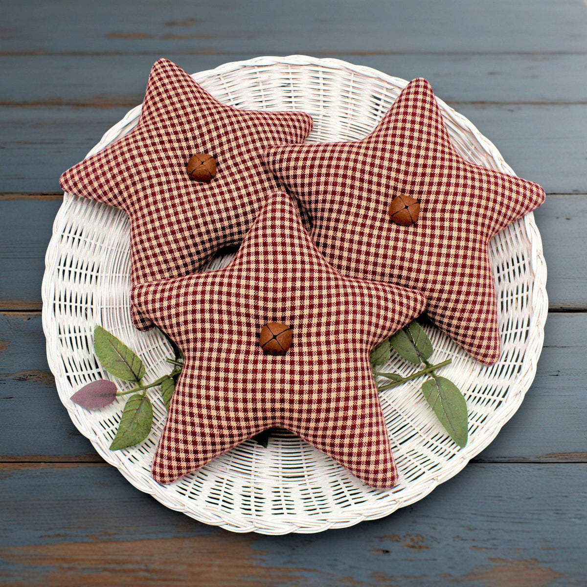 Three star fabric bowl fillers in a burgundy and beige check pattern, each with a rusty bell in the center, arranged on a white wicker plate over a blue wooden surface.