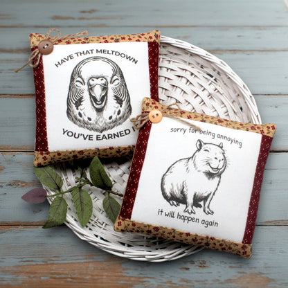 Two decorative fabric mini pillows displayed on a white woven tray over a rustic blue wooden surface. One pillow features a parakeet illustration with the text 'HAVE THAT MELTDOWN YOU'VE EARNED IT' and the other features a capybara illustration with the text 'sorry for being annoying it will happen again'.