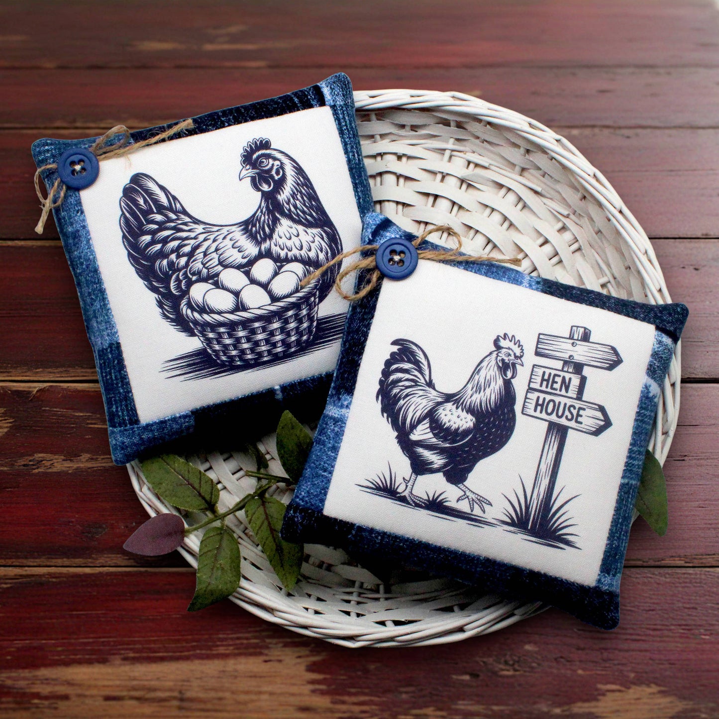 Two square decorative pillows with denim blue borders and white center panels featuring black chicken illustrations, displayed on a white woven tray over a dark wooden surface.