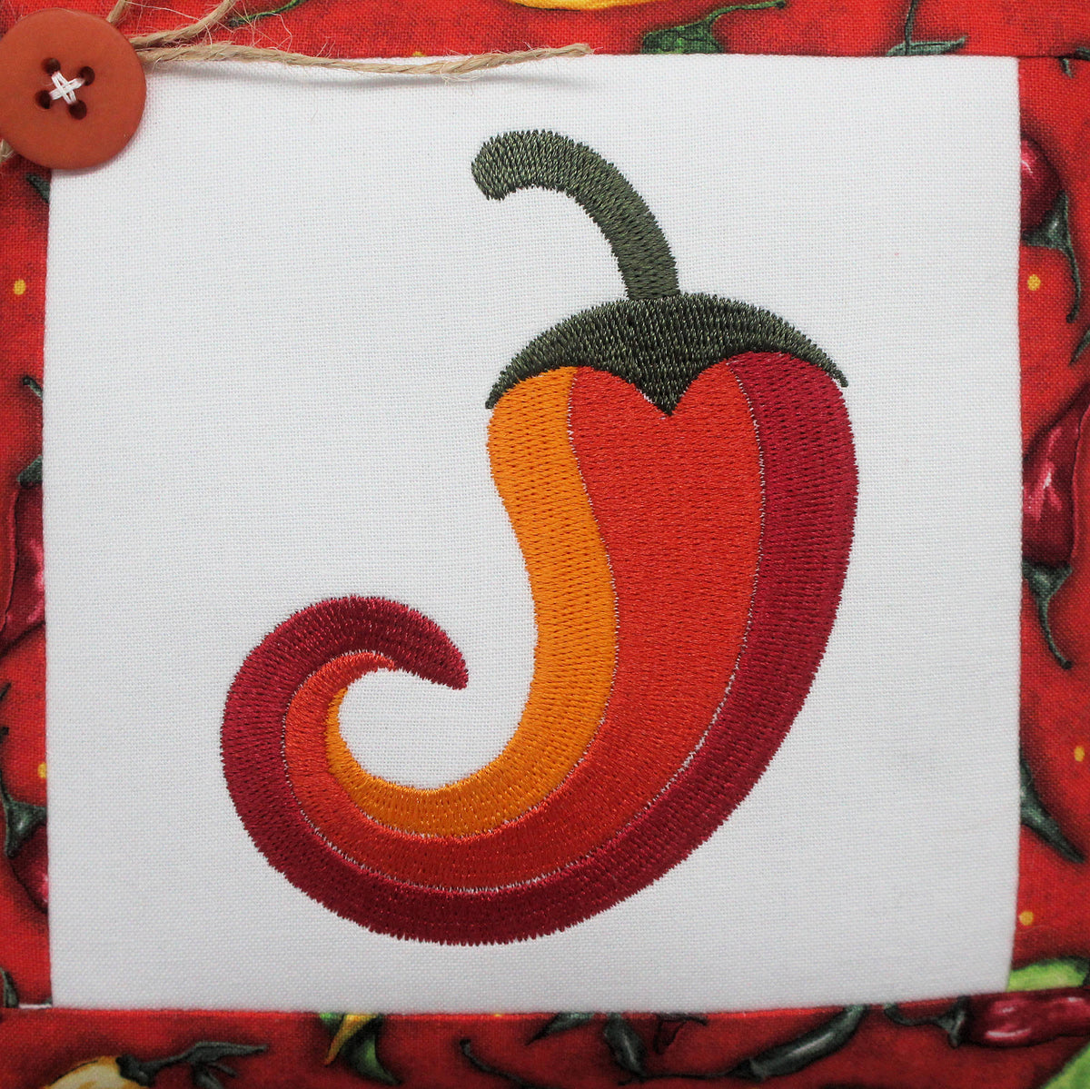 Embroidered chili pepper mini pillow tuck on white fabric square, surrounded by red patterned fabric with a brown button at the top left.