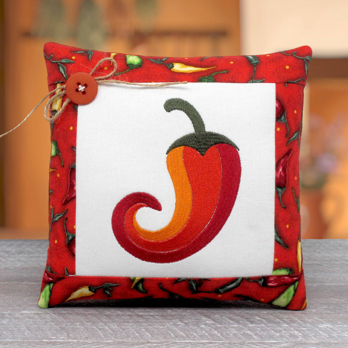A square decorative throw pillow with a red chili pepper border and a central white panel featuring an embroidered red and orange chili pepper, set on a gray surface with a blurred warm-toned background.
