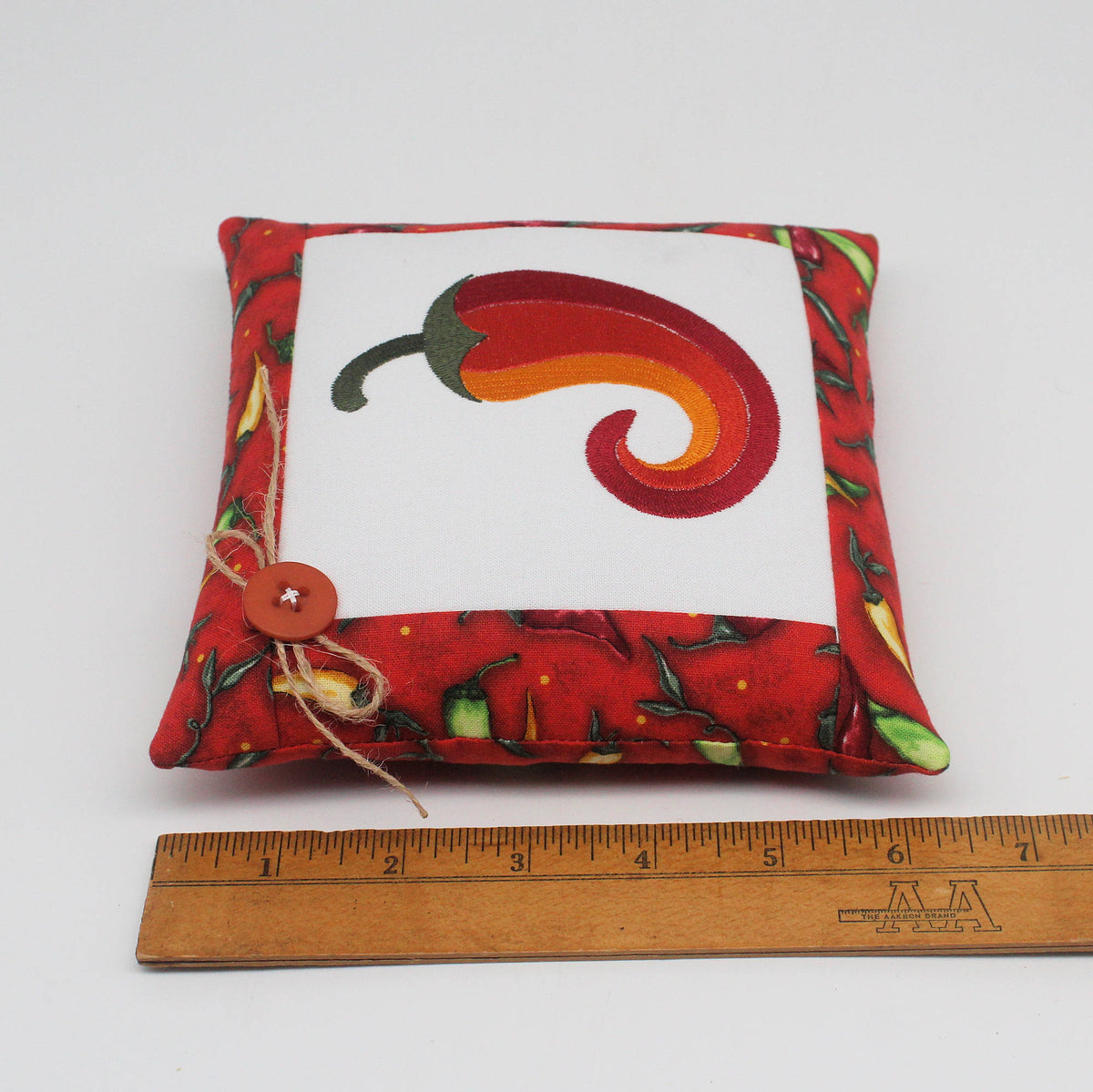 A square fabric pillow with a red chili pepper design on a white center panel, surrounded by a red chili-patterned border. A wooden ruler is placed below the pillow to show its size.