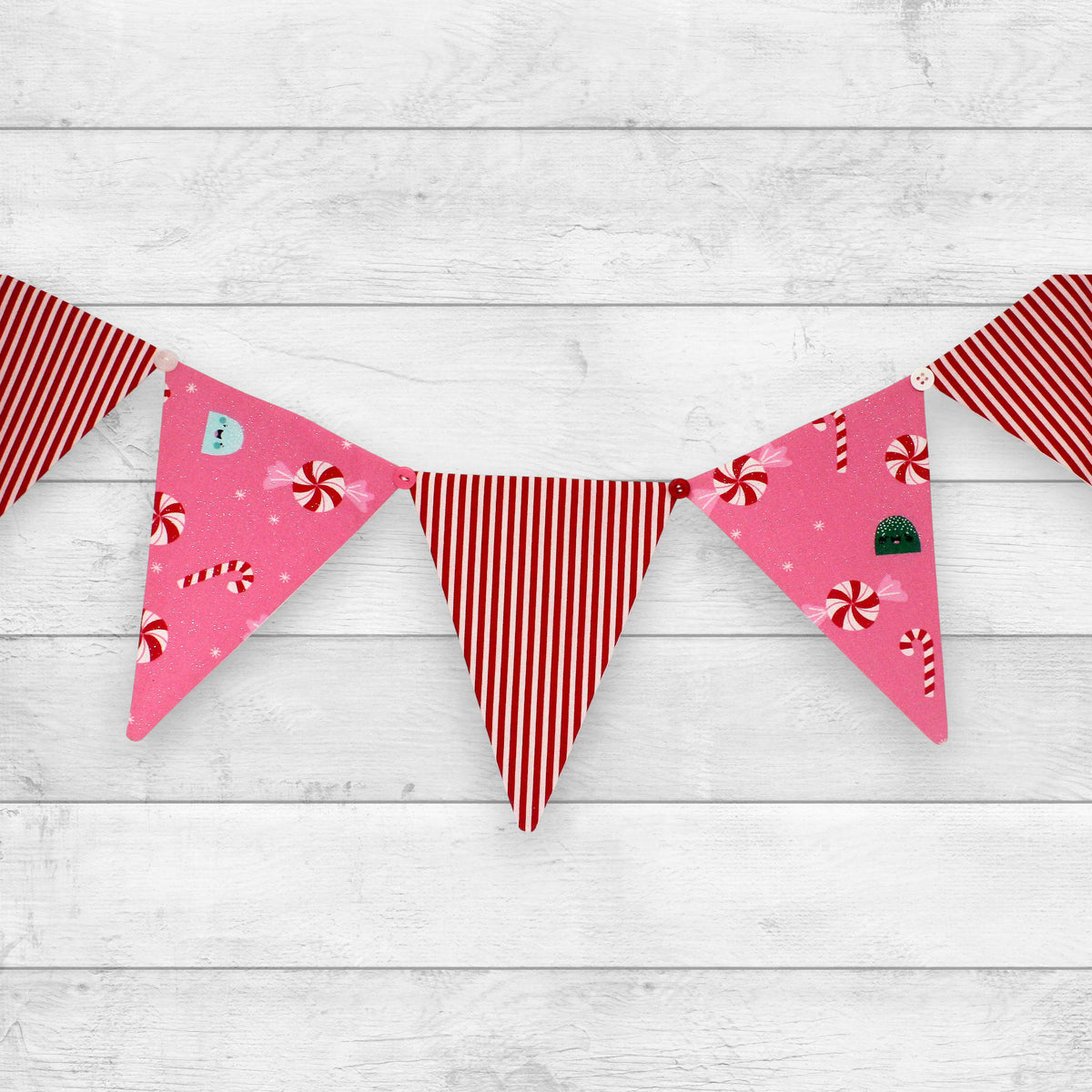 A festive bunting banner with triangular pennants hanging across a white wood plank background. The pennants feature holiday patterns including red and white stripes, pink backgrounds with red candy canes and peppermint candies, and one pink pennant with a small green Christmas tree.