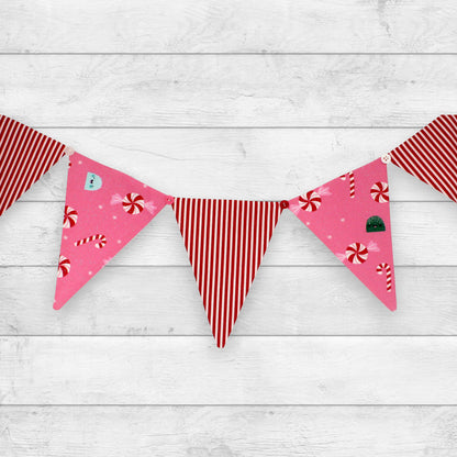 A festive bunting banner with triangular pennants hanging across a white wood plank background. The pennants feature holiday patterns including red and white stripes, pink backgrounds with red candy canes and peppermint candies, and one pink pennant with a small green Christmas tree.