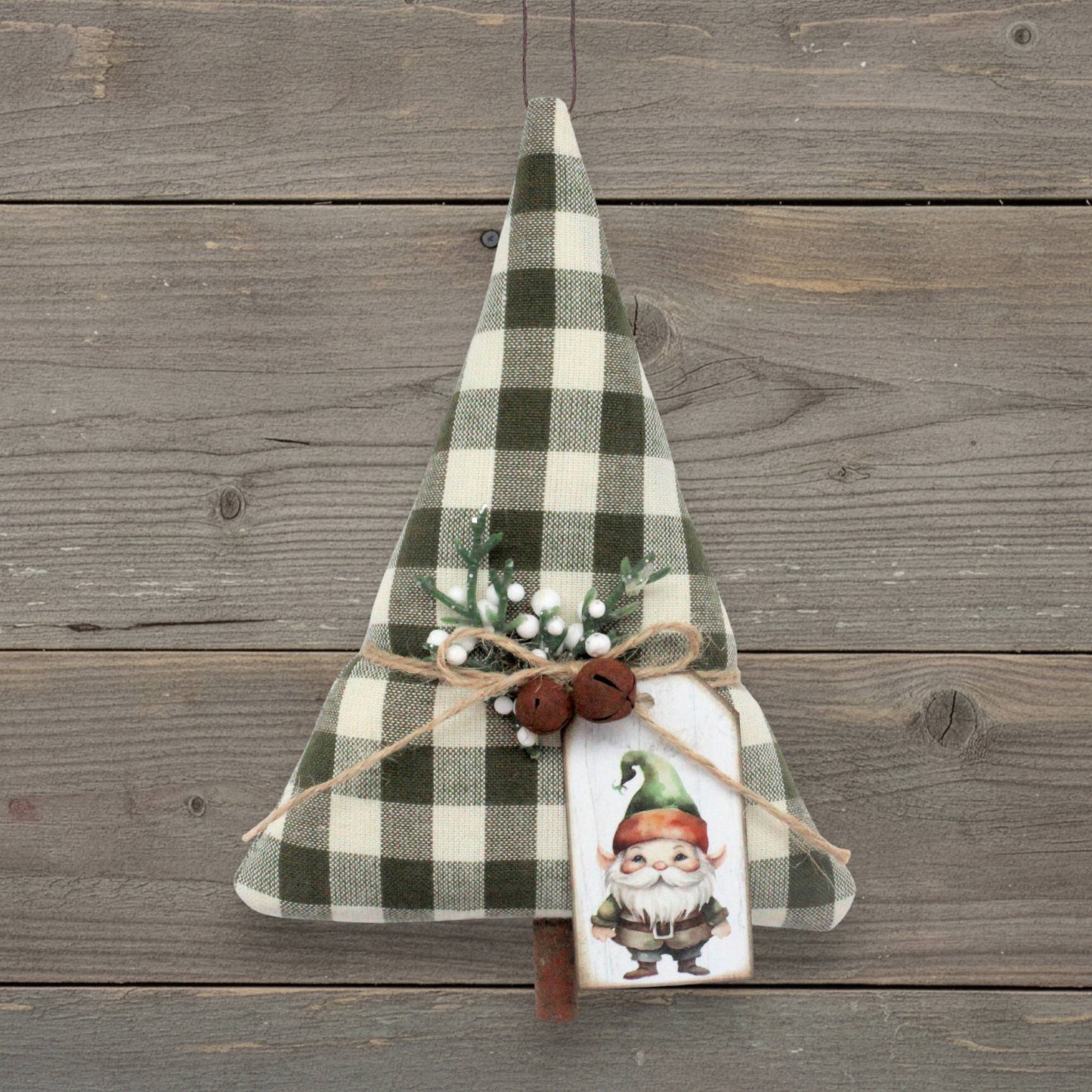 A rustic Christmas tree-shaped ornament made from green and cream buffalo plaid fabric, decorated with a jute twine bow, two small rusty bells, faux evergreen branches with white berries, and a painted gnome tag, displayed against a weathered gray wood plank background.