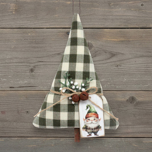 A rustic Christmas tree-shaped ornament made from green and cream buffalo plaid fabric, decorated with a jute twine bow, two small rusty bells, faux evergreen branches with white berries, and a painted gnome tag, displayed against a weathered gray wood plank background.