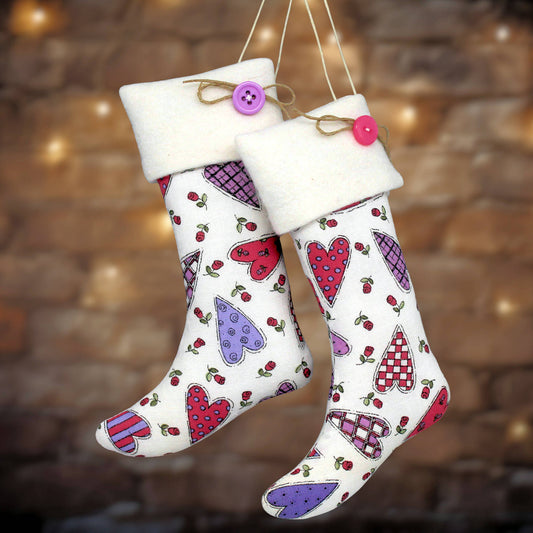 Two white Christmas stockings with a colorful heart pattern, hanging against a blurred warm brick wall background.