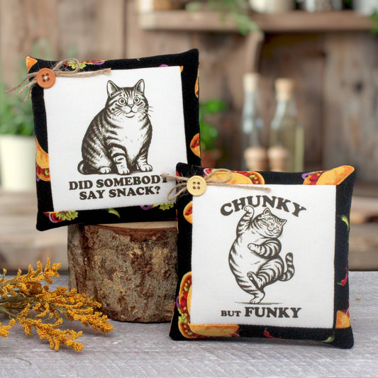 Two decorative square fabric mini pillows with black cat illustrations and humorous text, displayed on a rustic wood stump with blurred bottles and greenery in the background.