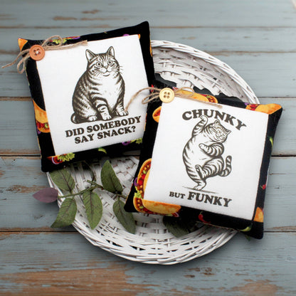 Two decorative square fabric mini pillows displayed on a white wicker tray over a distressed blue-gray wooden surface. Each pillow has a black border, a white center panel, a small wooden button, and a colorful patterned edge.