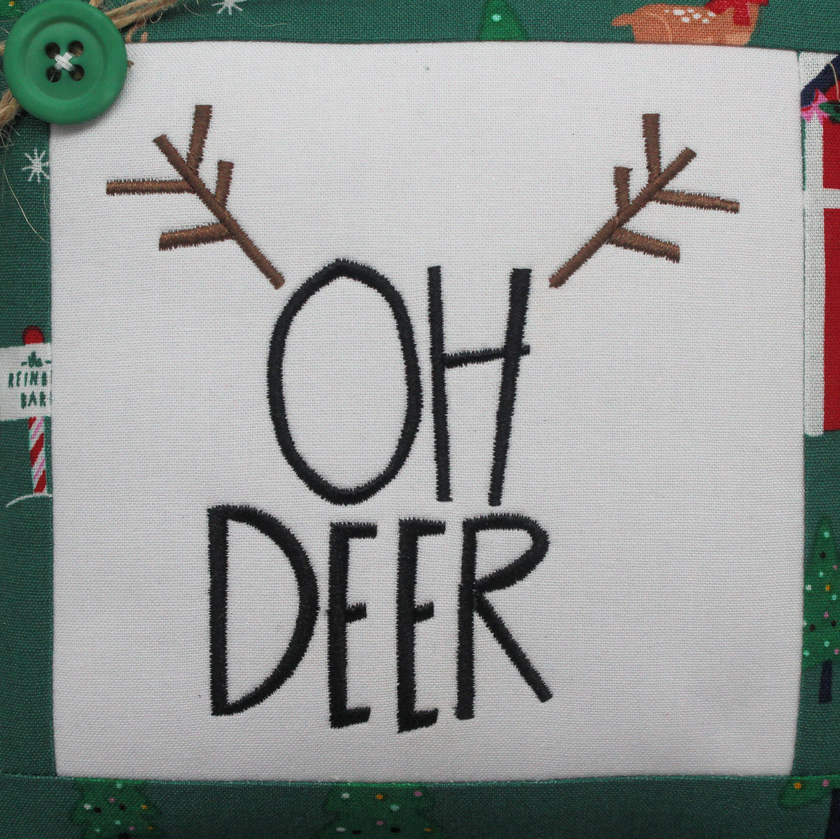 A square white fabric mini pillow with embroidered black text reading 'OH DEER' and brown antler motifs above, shown on a green holiday-themed fabric background with a green button and Christmas motifs.