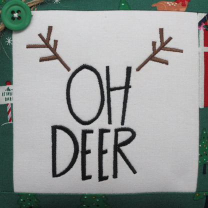 A square white fabric mini pillow with embroidered black text reading 'OH DEER' and brown antler motifs above, shown on a green holiday-themed fabric background with a green button and Christmas motifs.