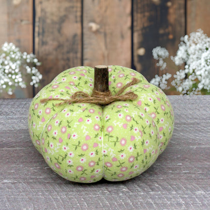 A fabric decorative pumpkin in light green with a pink and white floral print, tied with brown twine and featuring a faux wooden stem, displayed on a rustic gray wooden surface with a weathered wood plank background and white flowers in the distance.