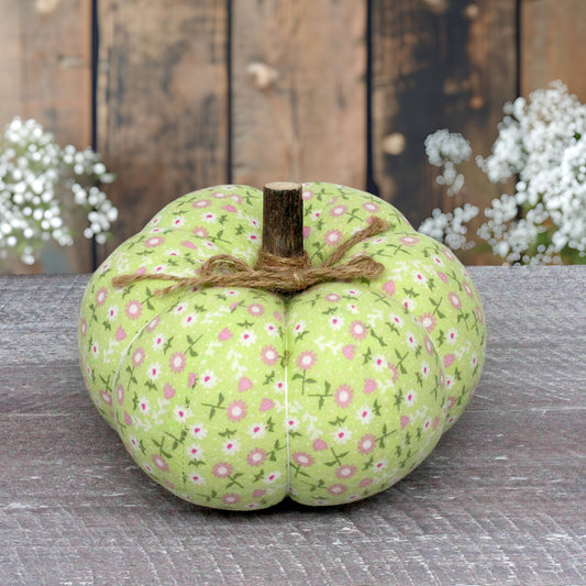 A fabric decorative pumpkin in light green with a pink and white floral print, tied with brown twine and featuring a faux wooden stem, displayed on a rustic gray wooden surface with a weathered wood plank background and white flowers in the distance.