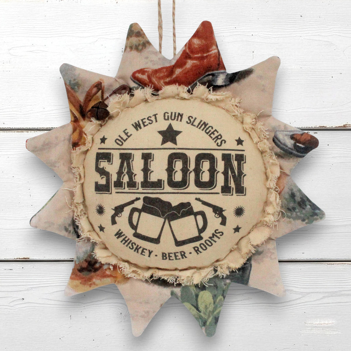 A rustic sunflower-shaped fabric wall hanging with a central circular patch reading 'OLE WEST GUN SLINGERS SALOON' and 'WHISKEY - BEER - ROOMS,' surrounded by distressed patchwork fabric in muted earthy tones.
