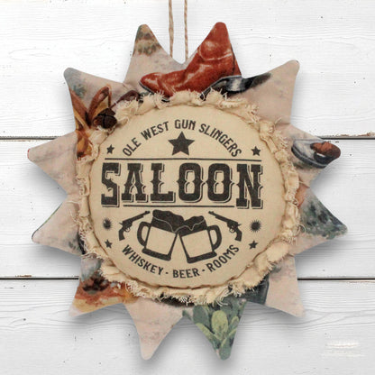 A rustic sunflower-shaped fabric wall hanging with a central circular patch reading 'OLE WEST GUN SLINGERS SALOON' and 'WHISKEY - BEER - ROOMS,' surrounded by distressed patchwork fabric in muted earthy tones.