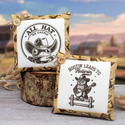 Two decorative square throw pillows with a tan and brown western-style border and white front panels, styled on a rustic wood stump with a blurred outdoor landscape background. One pillow shows a frog wearing a cowboy hat with the text 'ALL HAT NO CATTLE.