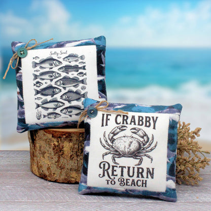 Two decorative square mini pillows with nautical designs are displayed on a rustic wooden stump against a beach backdrop. One pillow features a black-and-white fish illustration with the text 'Salty Soul,' and the other features a crab illustration with the phrase 'IF CRABBY RETURN TO BEACH.