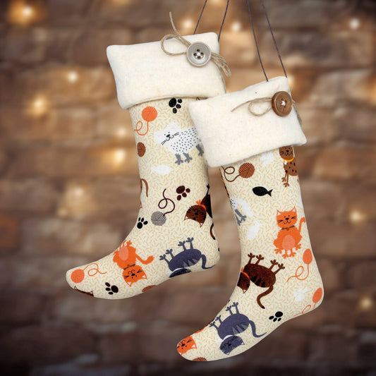 Two hanging Christmas stockings with cream fleece cuffs and a playful cat-themed print on a light beige background. The print features orange, brown, and blue cats, paw prints, and small toys.