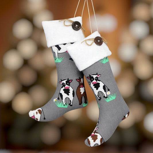 Two gray Christmas stockings with white cuffs, hanging by strings. The stockings are printed with cute cows set against a warm, blurred holiday background.