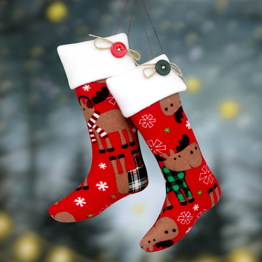 Two red Christmas stockings with white fleece cuffs hang against a blurred holiday background. One stocking features a moose wearing a red-and-white striped sweater, and the other features a moose wearing a green plaid sweater.