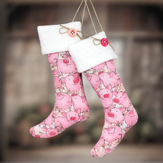 Two pink Christmas stockings with a cartoon pig print, white fleece cuffs, and hanging twine with decorative buttons, shown against a blurred rustic background.