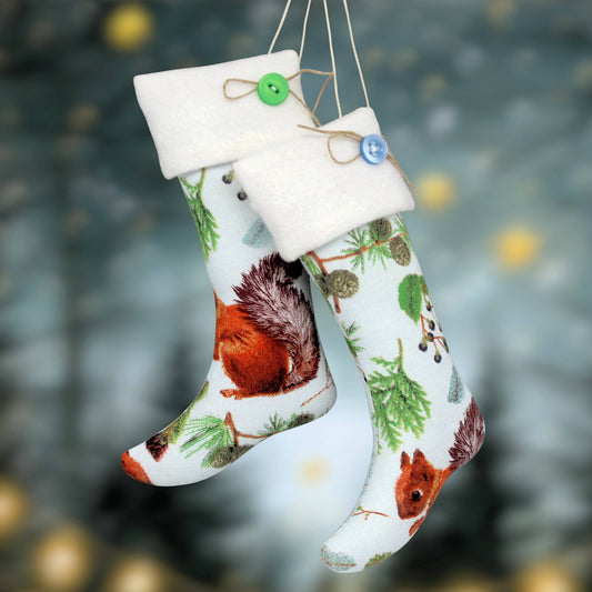 Two hanging Christmas stockings with white fleece cuffs, decorated with red squirrels, pine trees, and green leaves on a light background. One stocking has a green button and the other has a blue button, both with jute hanging strings against a blurred festive background.