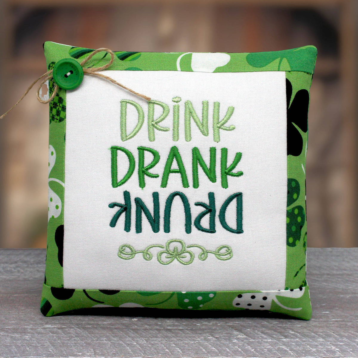 Small pillow tuck or bowl filler is embroidered with the phrase 'drink drank drunk' with the word 'drunk' upside down.  The center panel is trimmed with fabric with a shamrock design on a green background.