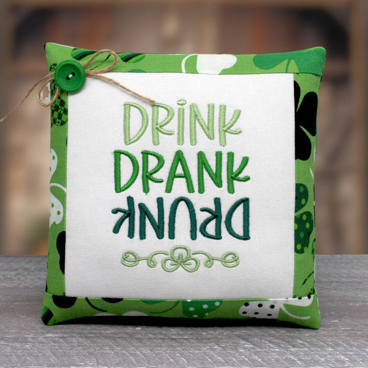 Small pillow tuck or bowl filler is embroidered with the phrase 'drink drank drunk' with the word 'drunk' upside down.  The center panel is trimmed with fabric with a shamrock design on a green background.
