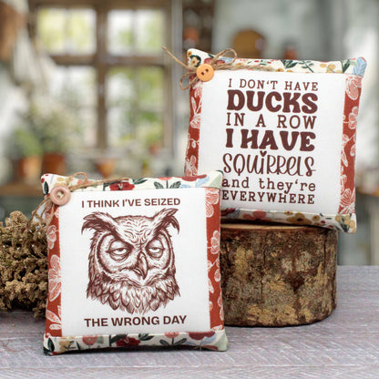 Two decorative fabric mini pillow-style signs with printed text and illustrations, styled on a tabletop with a rustic wood stump and blurred indoor background.