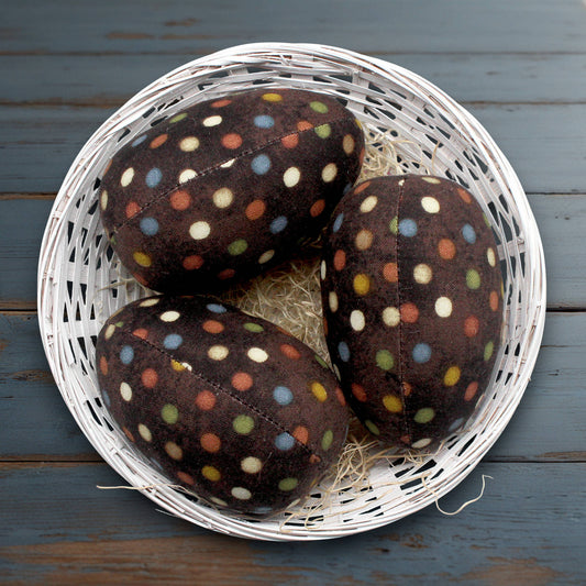 Three brown fabric eggs with colorful polka dots in a white basket on a wooden surface.