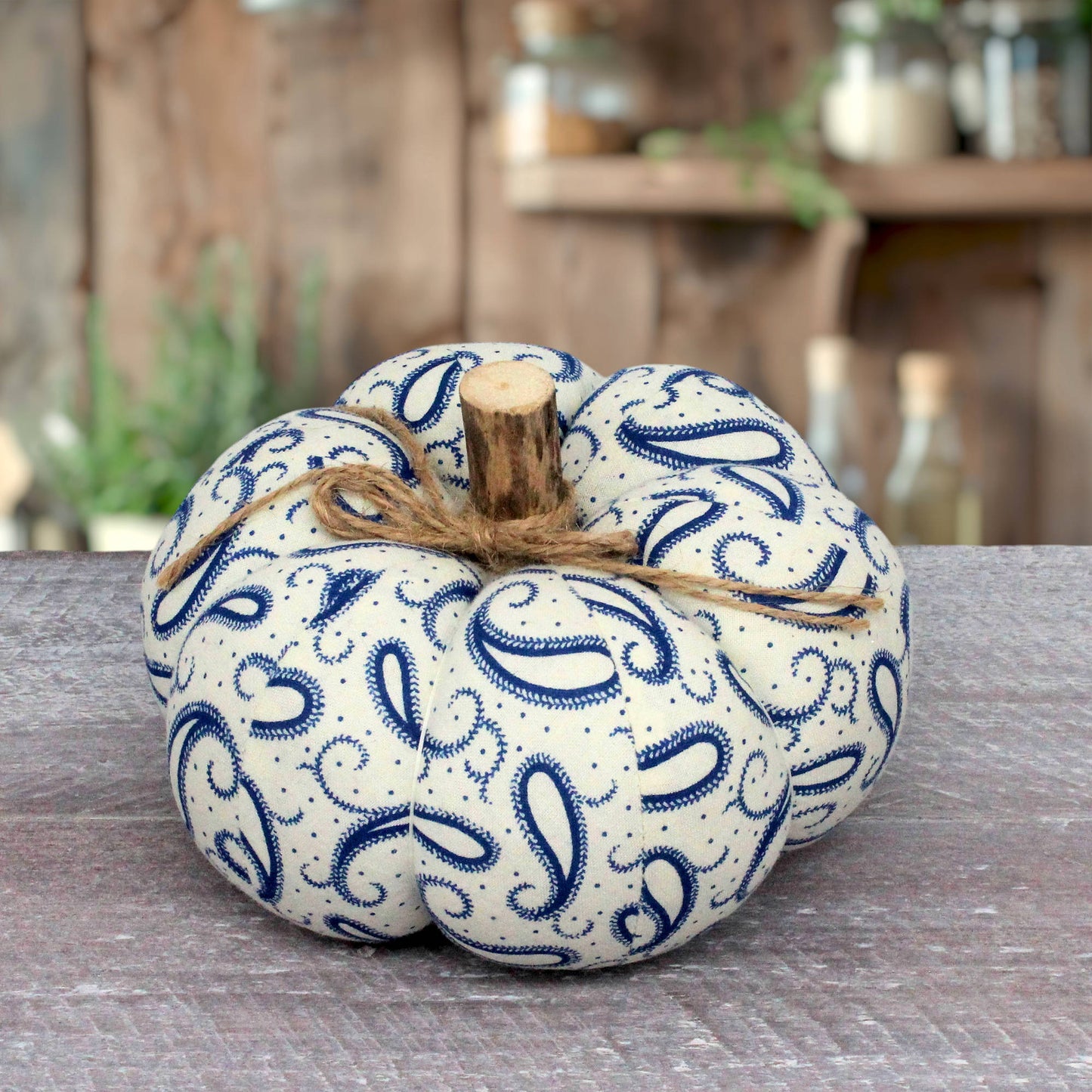 A fabric decorative pumpkin with a blue paisley pattern on a cream background, tied with natural twine and a small wooden stem, displayed on a rustic wooden surface.