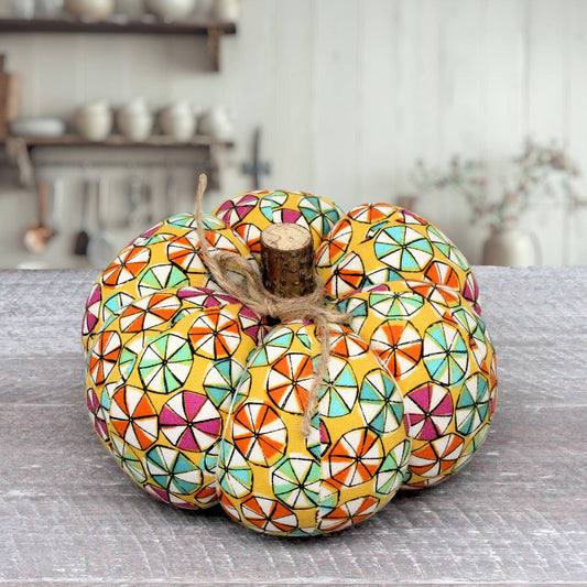 A decorative pumpkin with a colorful geometric candy pattern in yellow, orange, green, pink, and white, tied with twine and set on a gray tabletop in a softly blurred indoor setting.