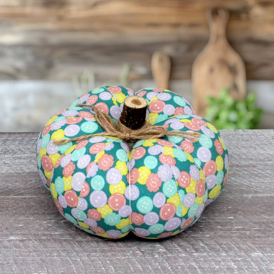 A fabric pumpkin decoration with a colorful button pattern in teal, pink, yellow, and lavender, tied with twine and topped with a small faux wood stem, displayed on a rustic gray wooden surface.