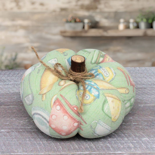 A fabric decorative pumpkin with a light green background and a multicolor kitchen-themed print, tied with twine and topped with a small wooden stem, displayed on a gray wooden surface.