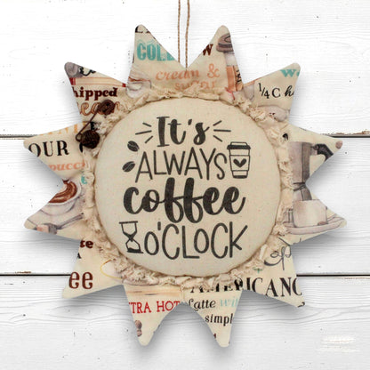 A sunflower-shaped decorative wall hanging with a central round disc that reads “It’s ALWAYS coffee o'clock,” surrounded by pointed rays made from vintage-style coffee-themed paper with a ruffled fabric trim.