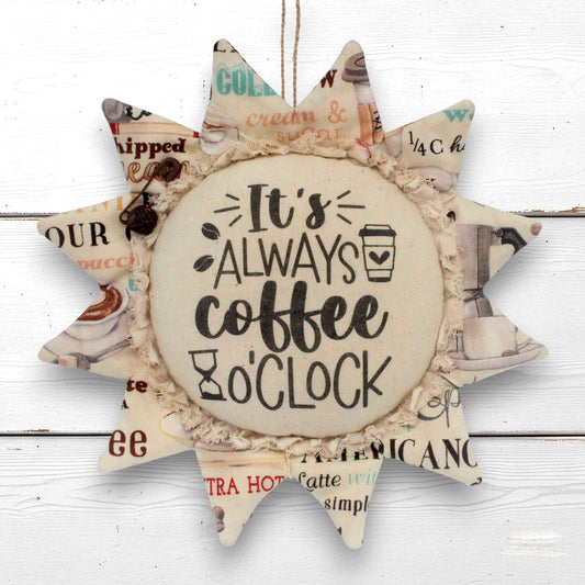 A sunflower-shaped decorative wall hanging with a central round disc that reads “It’s ALWAYS coffee o'clock,” surrounded by pointed rays made from vintage-style coffee-themed paper with a ruffled fabric trim.