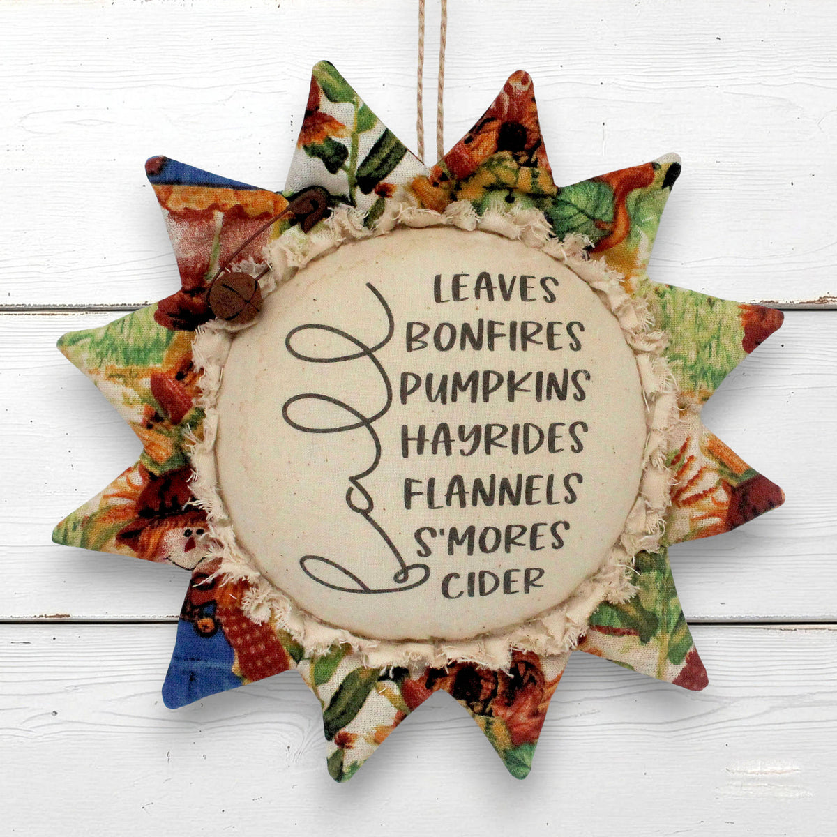 A fabric sunflower-shaped hanging ornament with a rustic printed patterned border and a central cream circle that reads, 'LEAVES BONFIRES PUMPKINS HAYRIDES FLANNELS S'MORES CIDER,' shown against a white wood plank background.