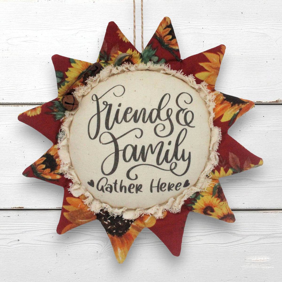 A fabric sunflower ornament wall hanging with the phrase “Friends & Family Gather Here” in black script on a cream center panel, surrounded by red and autumn floral rays on a white wood plank background.