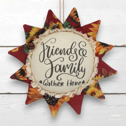 A fabric sunflower ornament wall hanging with the phrase “Friends & Family Gather Here” in black script on a cream center panel, surrounded by red and autumn floral rays on a white wood plank background.