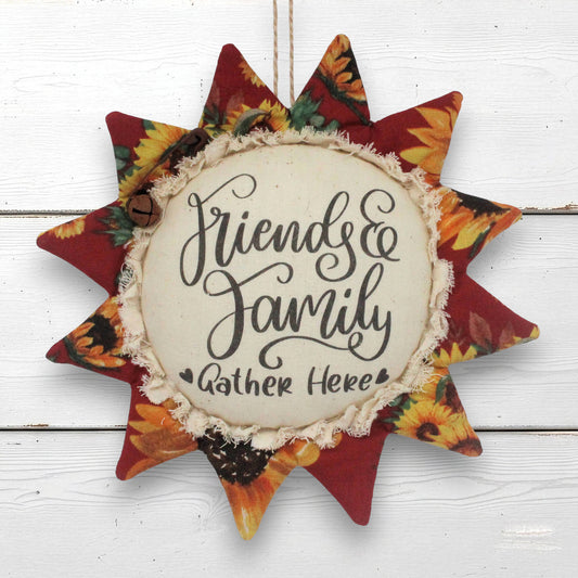 A fabric sunflower ornament wall hanging with the phrase “Friends & Family Gather Here” in black script on a cream center panel, surrounded by red and autumn floral rays on a white wood plank background.