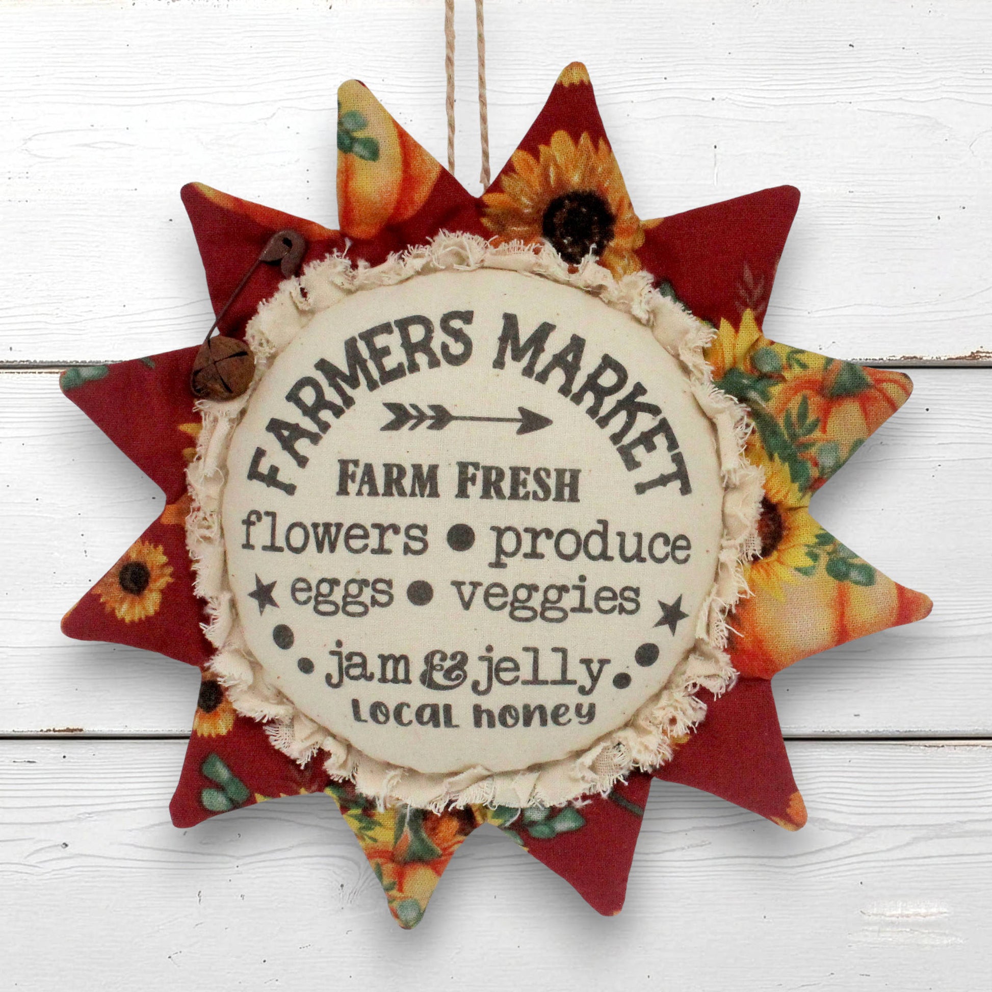 A fabric sunflower-shaped hanging sign with a central cream circle reading 'Farmers Market' and autumn-themed printed fabric points in red, orange, yellow, and floral patterns, shown against a white wood plank background.