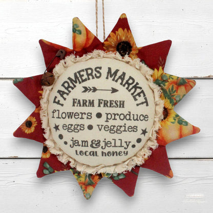 A fabric sunflower-shaped hanging sign with a central cream circle reading 'Farmers Market' and autumn-themed printed fabric points in red, orange, yellow, and floral patterns, shown against a white wood plank background.