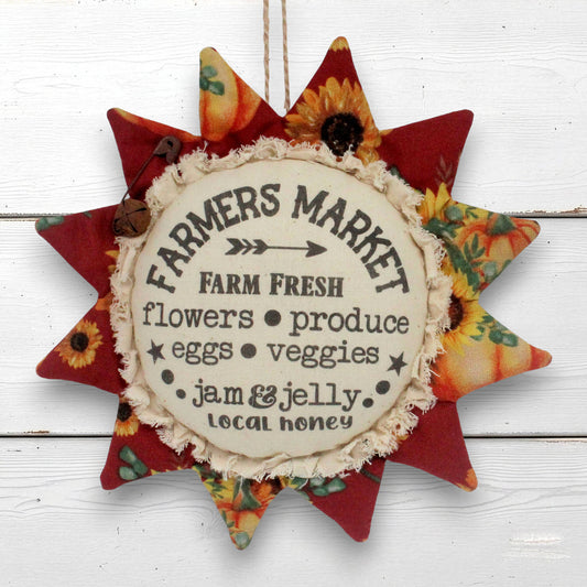 A fabric sunflower-shaped hanging sign with a central cream circle reading 'Farmers Market' and autumn-themed printed fabric points in red, orange, yellow, and floral patterns, shown against a white wood plank background.