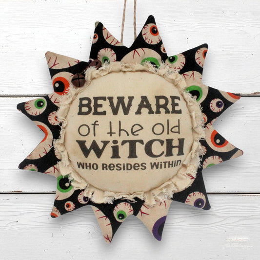 A sunflower-shaped fabric wall hanging with a central circular patch that reads 'BEWARE of the old WITCH WHO RESIDES WITHIN'. The outer points feature a black fabric pattern with embroidered-style eyes in green, white, and red.
