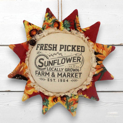 A fabric sunflower-shaped wall hanging ornament with a central cream circle reading 'FRESH PICKED Sunflower Farm & Market Locally Grown Est. 1904' and patterned star points in red, yellow, orange, and green, shown hanging against a white wood plank background.