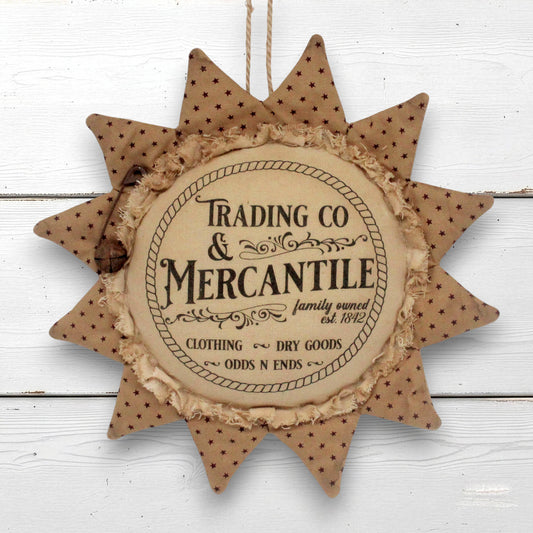A rustic sunflower-shaped hanging ornament with a central round plaque reading 'Trading Co & Mercantile' and decorative text around it, shown against a white painted wood background.