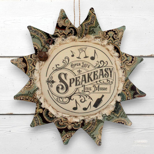 A rustic sunflower-shaped fabric wall hanging with a central cream circle reading 'Open Late by Speakeasy Live Music' and surrounded by patterned green, beige, and brown star points, hanging against a white wood plank background.