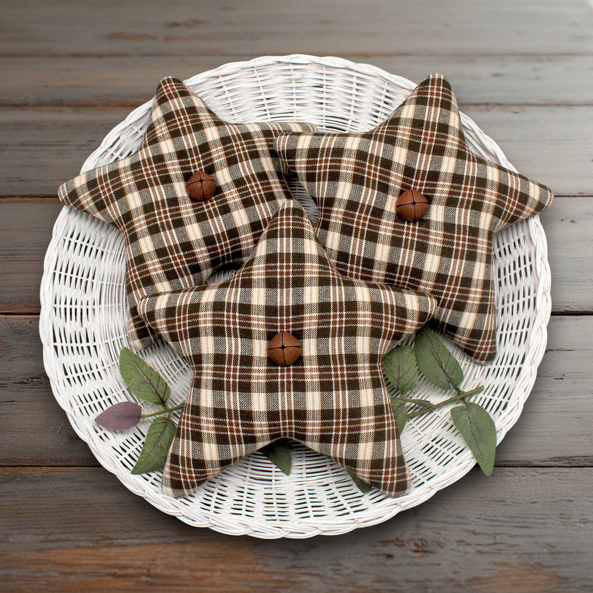 Three fabric homespun star bowl fillers in a brown and beige plaid pattern, each with a rusty bell in the center, arranged on a white wicker tray over a rustic wooden surface.